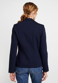 Marynarka w kolorze granatowym o dopasowanym kroju, z długimi rękawami i detalami na szwie z tyłu. Gładki materiał i usztywnione klapy zapewniają wyszczuplający wygląd.
