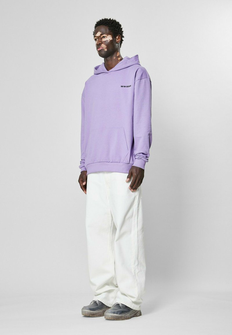Sudadera con capucha púrpura y bolsillo frontal, combinada con pantalones blancos holgados y calzado gris. Detalle de capucha ajustable visible.