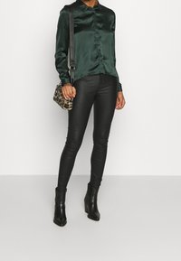 Dunkelgrünes Satinhemd mit Knöpfen, kombiniert mit schwarzen Kunstleder-Leggings und schwarzen Absatzstiefeletten. Die Tasche hat ein Leopardenmuster.