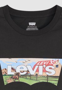 Svart bomull t-skjorte med et vintage Levi's-logo, landskapsmotiv med en hest, låve, gress og fugler, rammet inn av en fargerik grafikk.