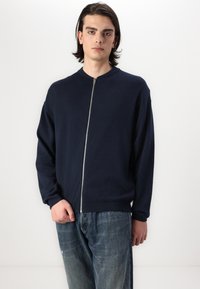 Sweater azul marinho com fecho éclair e gola redonda, com punhos ribateados. Feito de material de malha suave, conta com um ziper frontal de comprimento total.