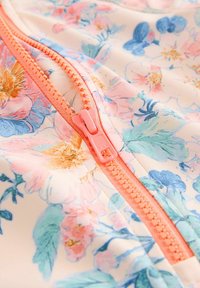 Blomstrede zip-up beklædning med pastelfarvede blomster i pink og blå på en lys baggrund, med en koralfarvet lynlås og blød stofstruktur.