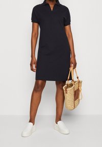 Vestido polo navy con mangas cortas y cuello, hecho de algodón. Combinado con zapatillas blancas sin cordones y un bolso de mimbre con detalles de cuero.