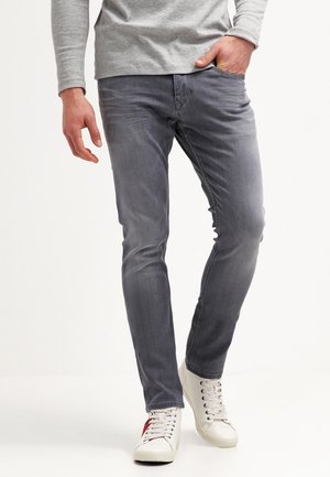 Uomo con jeans grigi aderenti, maglia a maniche lunghe grigio chiaro e sneakers bianche con dettagli rossi e neri, in piedi con una mano in tasca.