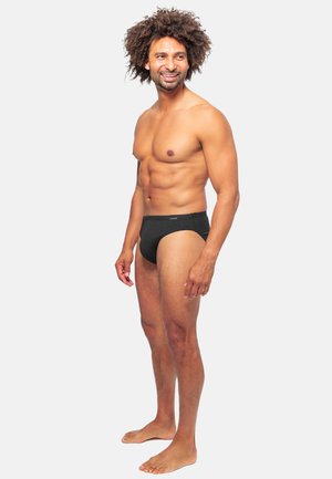 Schwarze Schwimmslips aus strukturiertem Material, mit einem niedrigen Bund und einem elastischen Taillenbund, getragen von einem barfüßigen Mann.