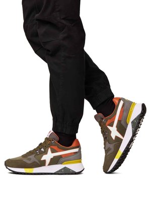 Personne portant un pantalon noir et des baskets marron, orange et jaune avançant sur un fond blanc.