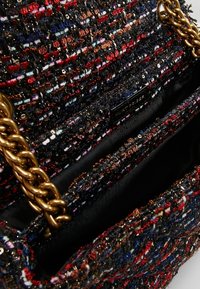 Detailní záběr na texturovanou vícebarevnou kabelku z tkaniny tweed se zlatým řetízkem a černou podšívkou.
