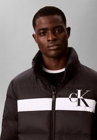 Jeune homme portant une doudoune noire avec une bande horizontale blanche et le logo "cK" sur la poitrine, debout devant un fond gris.