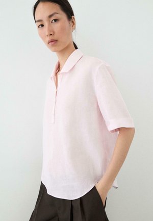 Kobieta ubrana w bladoróżową koszulę z krótkim rękawem z kołnierzykiem i ciemne spodnie, stojąca z jedną ręką w kieszeni na jednolitym jasnym tle.