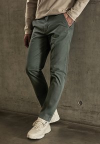 Pantalons en toile de coton verts avec une coupe slim, poches avant et détails de couture discrets, associés à des chaussures de sport blanches.