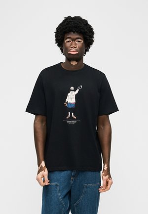 Άνδρας με λεύκη που φοράει μαύρο T-shirt με γραφικό στοιχείο μιας φιγούρας που κρατά χειροπέδες και μπλε τζιν, στέκεται μπροστά σε μονόχρωμο φόντο.