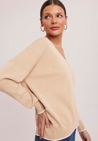 Beżowy sweter dzianinowy z dekoltem w serek, z białym wykończeniem na dekolcie i mankietach. Długie, luźne rękawy zwiększają komfort.