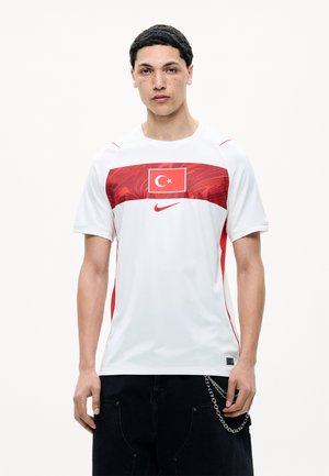 Ung mand iført en hvid og rød sportstrøje med det tyrkiske flag og Nike-logo, kombineret med sorte bukser og et kædeaccessory.