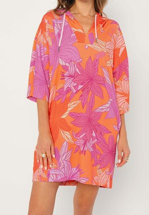 Kobieta ubrana w luźną mini sukienkę z kapturem, ozdobioną dużymi kwiatowymi wzorami w kolorach pomarańczowym, różowym i fioletowym oraz rękawami o długości do łokcia.