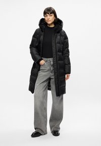 Piumino nero con cappuccio, chiusura zip e design trapuntato. Indossato sopra una maglietta nera e jeans grigi larghi, che mostrano uno stile casual.