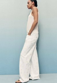 Pantalon large blanc avec broderie florale dorée associé à un débardeur léger, présenté sur un fond dégradé bleu.