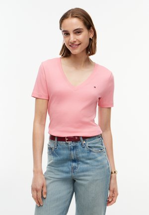 Frau lächelt, trägt ein rosa kurzärmeliges V-Ausschnitt-Shirt, das in hellblaue Jeans mit rotem Gürtel gesteckt ist, vor weißem Hintergrund.