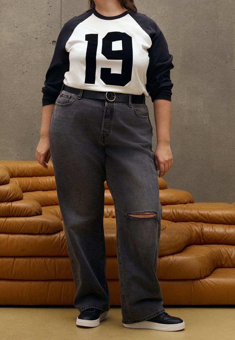 T-shirt raglan blanc et noir avec l'impression "19", associé à un jean gris ample avec une déchirure. Ceinture noire avec une boucle ronde, et baskets noires.