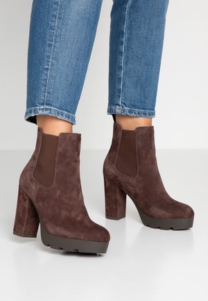 Bottines à plateforme en daim marron avec talons épais portées avec un jean en denim bleu sur un sol blanc.