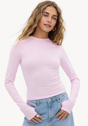 Vrouw met golvend blond haar, gouden creolen, een nauwsluitend lichtroze shirt met lange mouwen, een blauwe spijkerbroek en meerdere ringen aan haar vingers.