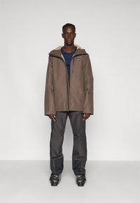 Giacca impermeabile marrone con fodera beige, zip frontale, due tasche laterali e cappuccio regolabile, abbinata a pantaloni grigio scuro e stivali.