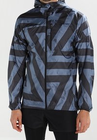 Veste légère à capuche de couleur noire avec des motifs géométriques bleus. Comprend une poche poitrine zippée et des poignets élastiques pour un ajustement sécurisé.