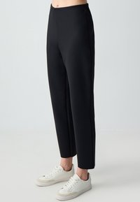 Pantalons noirs ajustés à taille haute, fabriqués en tissu lisse, avec une coupe droite. Associés à des baskets blanches.