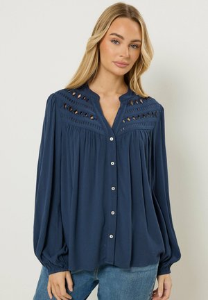 Blondynka w luźnej, granatowej koszuli na guziki z koronkowymi wycięciami na ramionach i długimi bufkami na rękawach, w połączeniu z niebieskimi dżinsami.