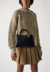 Borsa a tracolla nera trapuntata con doppi manici, tracolla a catena e dettaglio del logo, abbinata a un maglione a righe e una gonna plissettata beige.