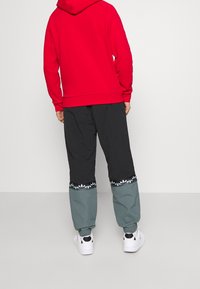 Červená mikina s kapucí v kombinaci s černými a zelenými kalhotami s květinovým vzorem na lemu a bílými sportovními botami. Celkově hladká textura.