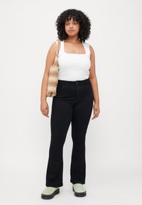 Débardeur blanc à encolure carrée, jeans noirs évasés, chaussures vertes à enfiler et un sac en bandoulière beige en tissu tissé. Cheveux frisés et foncés encadrant le visage.