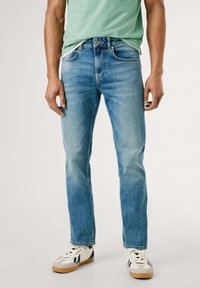 Jeans de mezclilla azul claro con un corte recto, que cuentan con cinco bolsillos, un ligero desgaste y un cierre de botones. Combinados con zapatillas blancas y negras.