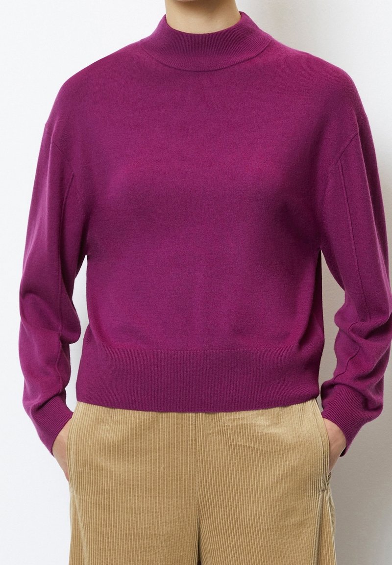 Personne portant un pull magenta à manches longues avec un col montant et un pantalon en velours côtelé beige, mains dans les poches, sur un fond uni.