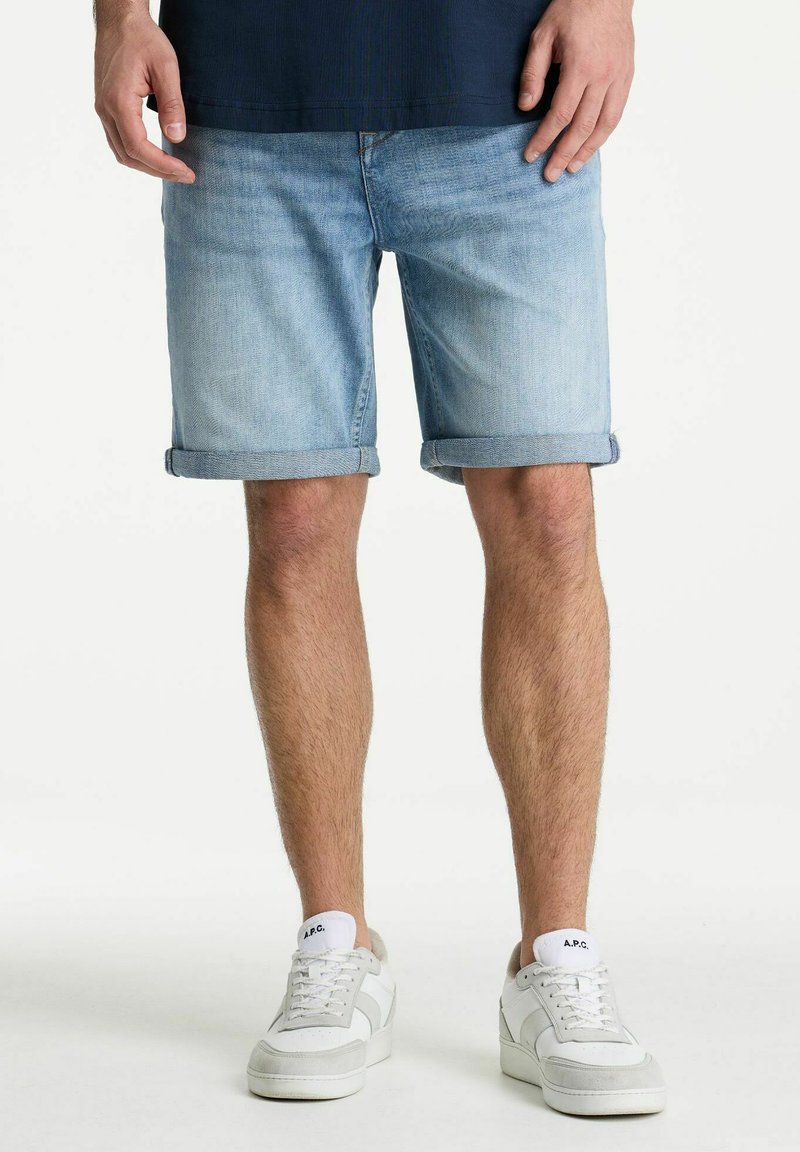 Man draagt lichtblauwe opgerolde spijkershorts en witte sneakers en staat tegen een effen achtergrond.