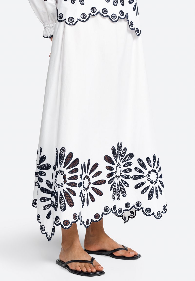 White maxi skirt with navy embroidery featuring floral patterns and scalloped hem. Paired with black flip-flops. Lightweight cotton fabric.