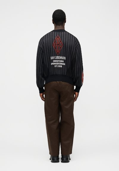 Black and grey striped sweater with oversized sleeves, featuring red embroidered logo and text on the back. Brown trousers and black footwear.