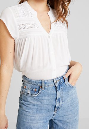 Femme portant un chemisier blanc à manches courtes avec des détails en dentelle et un jean en denim bleu clair taille haute, main reposant dans la poche.