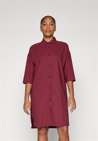 Vestido camisa burdeos con mangas cortas, un frente abotonado y un corte relajado. Fabricado en un tejido suave con sutil textura y con aberturas laterales.