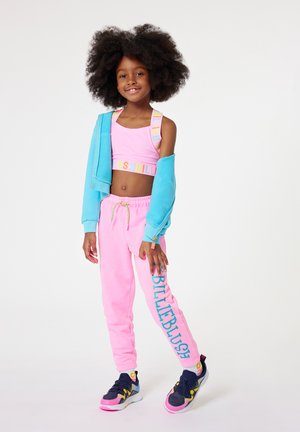 Top cropped rose avec le bandeau "BILLIEBLUSH", un pantalon de survêtement rose assorti et un sweat à capuche bleu zippé. Détails avec des accents colorés et des baskets.