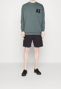 Grön tröja med svart "AX"-logotyp, parat med svarta cargoshorts. Modellen har på sig svarta slip-on skor och står mot en enkel bakgrund.