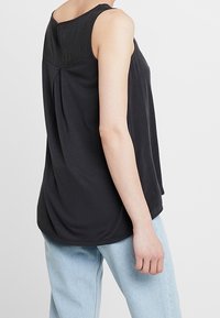 Top senza maniche nero con un design plissettato sul retro, caratterizzato da un collo rotondo e un tessuto morbido e liscio. Abbinato a jeans azzurri chiari.