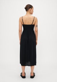 Μαύρο φόρεμα μεσαίου μήκους με ρυθμιζόμενους λεπτούς ιμάντες, πτυχές στην πίσω πλευρά και ρευστή φούστα. Φορεμένο με χοντρές μαύρες σανδάλια.