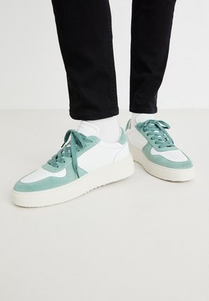 Personne portant des baskets blanches et vert clair avec des chaussettes blanches et un pantalon noir, debout sur un sol blanc uni.