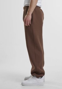 Un pantalon de jogging marron avec une coupe décontractée, une taille élastique, des poches latérales et des poignets côtelés, associé à des baskets blanches. Texture lisse.