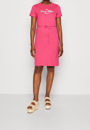 Femme portant une robe mi-longue rose Tommy Hilfiger avec une ceinture à la taille et des sandales compensées beiges sur un fond uni.