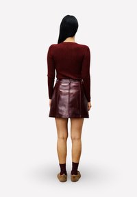 Vrouw met lang donker haar, gekleed in een geribbelde bordeauxrode trui, een bordeauxrode leren rok, donkere sokken en bruine slippers, gezien van achteren.