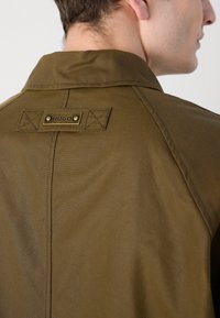 Olive green jacket with a pointed collar, featuring a branded metal tag and cross-stitched accents on the back. Smooth texture, tailored fit.