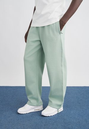 Persona con pantalones deportivos verde claro y zapatillas blancas de pie sobre un suelo alfombrado azul con una mano en el bolsillo.