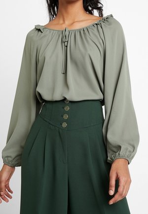 Femme portant un chemisier vert olive clair à épaules dénudées avec de longues manches bouffantes et un pantalon taille haute vert foncé avec quatre boutons.