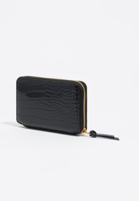 Cartera negra con estampado de cocodrilo hecha de material brillante. Cuenta con un cierre dorado y una lengüeta de tiro adjunta. Forma rectangular compacta.
